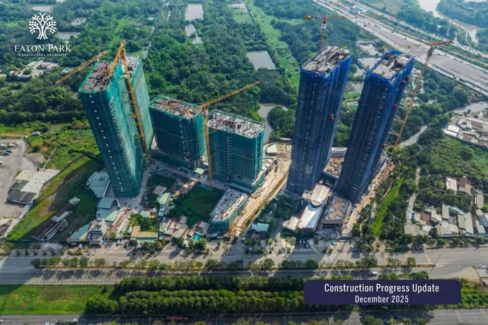 Aerial photograph of Eaton Park in December 2025. The towers are rising under green and blue safety netting with tower cranes in place, and the podium pools and landscaping are under construction between them.