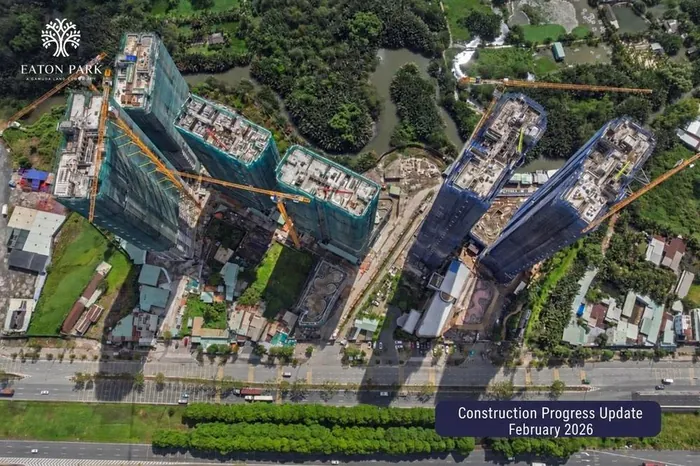 Overhead aerial photograph of the Eaton Park site in February 2026. The towers stand under green and blue safety netting with tower cranes in place, the podium pools and landscaping are taking shape between them, and Mai Chi Tho Boulevard runs along the bottom of the frame.