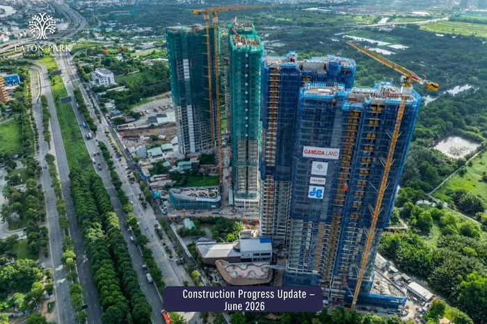 Aerial photograph of Eaton Park in June 2026. The towers stand at full height under green and blue safety netting with tower cranes still in place, contractor banners on the facade, and the podium and landscaping taking shape below.
