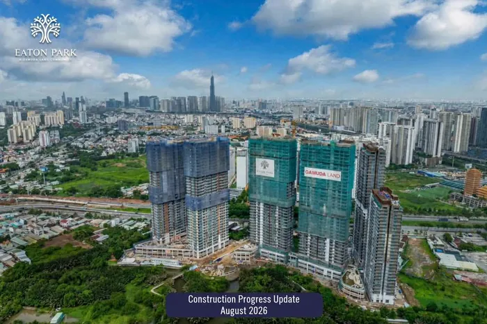 Aerial photograph of Eaton Park in August 2026. All six towers stand at full height with the safety netting coming off the lower floors, facade and balconies visible, the podium and landscaping largely formed, and the city skyline behind.
