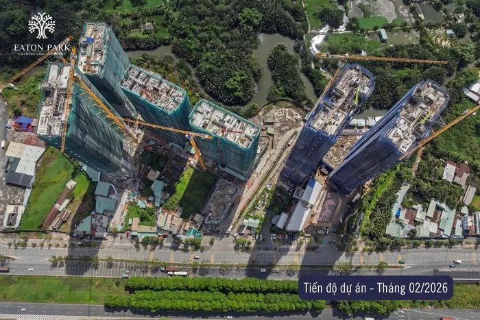 Ảnh chụp tiến độ Eaton Park thẳng từ trên xuống, tháng 2 năm 2026. Các tòa tháp dưới lưới bao che xanh lá và xanh dương, hồ bơi khối đế và cảnh quan đang hình thành, đại lộ Mai Chí Thọ chạy dọc mép dưới khung hình.